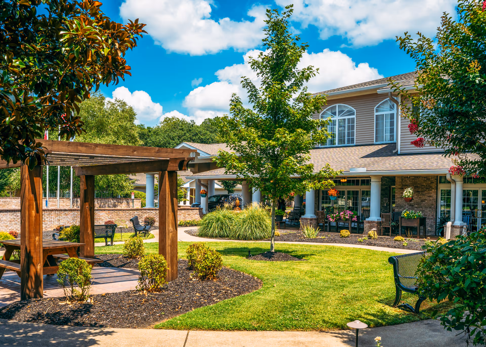 Outdoor garden area of a senior living facility with a wooden pergola over a picnic table, green grass, trees, shrubs, benches, and a two-story building with large windows and a covered porch with rocking chairs and hanging flower pots under a blue sky with clouds.