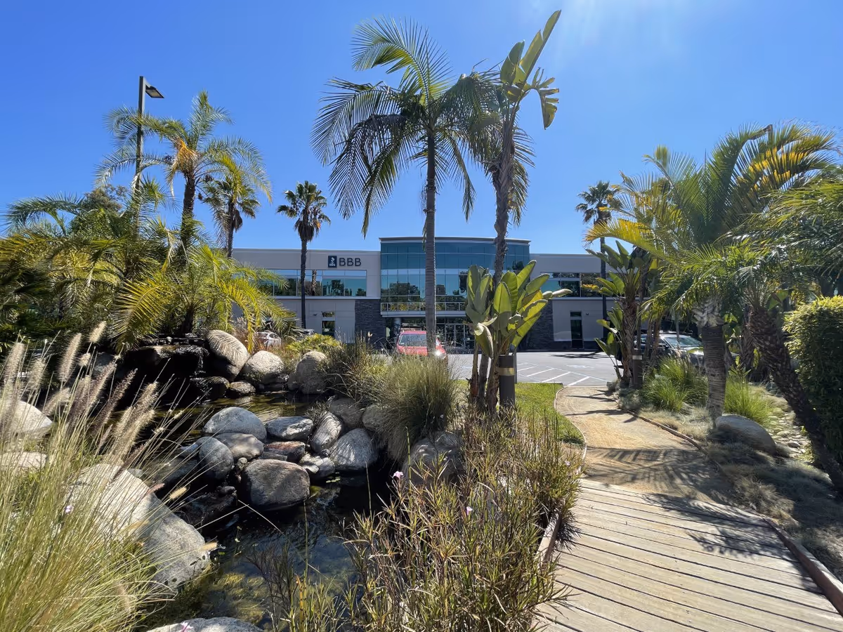 Landscaped entrance with palm trees, a rock water feature and a wooden walkway leading to a two-story glass-front building.