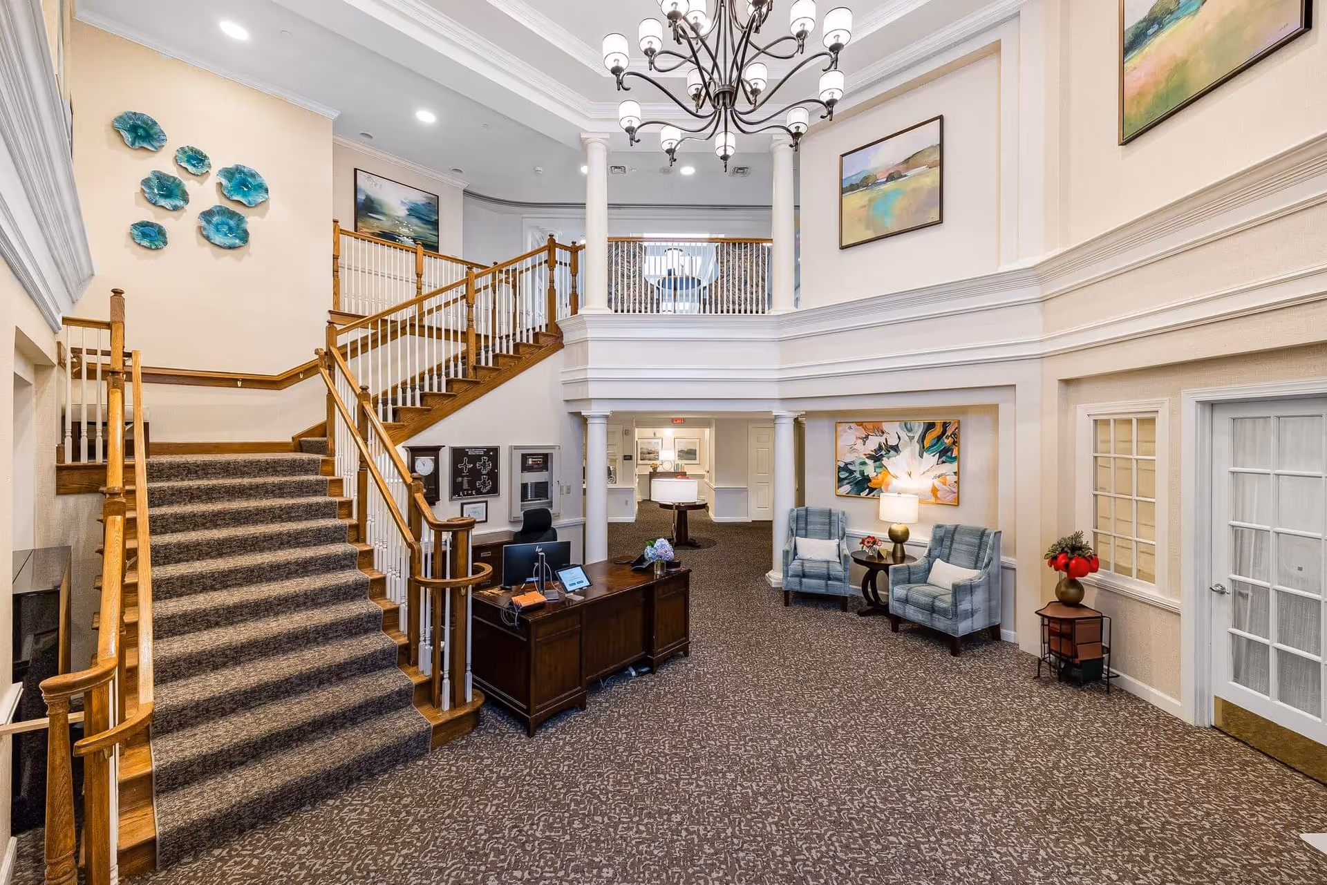 Elegant senior living lobby with staircase, chandelier, reception desk, seating area, and artwork.