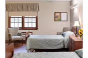 Two twin beds in a beige senior living room with a window and furnishings.