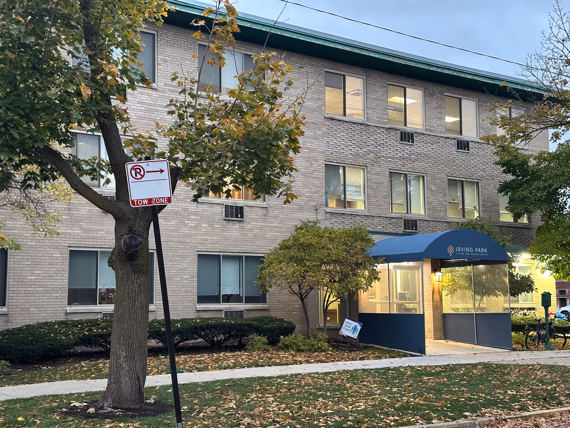 Exterior view of a three-story brick building with multiple windows, a blue awning entrance labeled 'Irving Park Living and Rehab Center,' surrounded by trees and bushes with fallen leaves on the ground.
