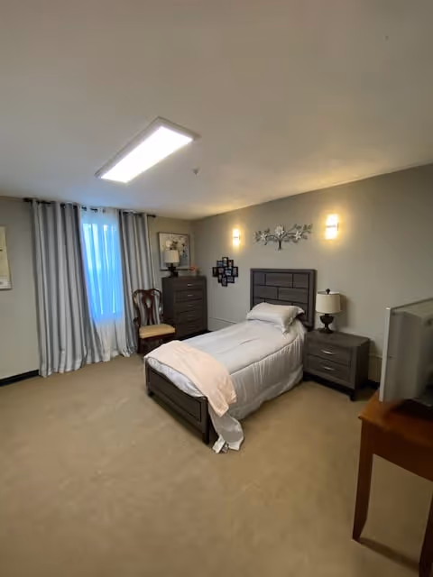 A neatly arranged bedroom with a single bed covered in white bedding and a light pink throw blanket. The room features a wooden headboard, a nightstand with a lamp, a dresser, a wooden chair, and a window with gray curtains. The walls are light-colored with two wall-mounted lights and decorative wall art above the bed.