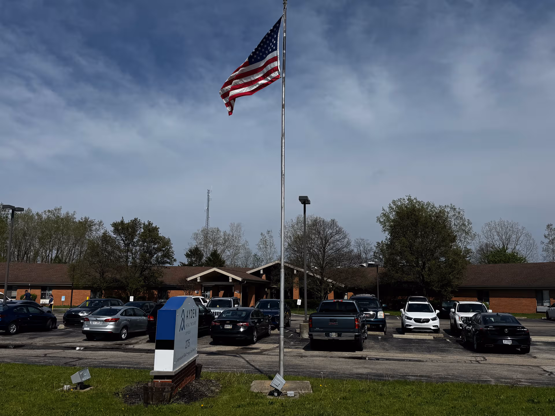American flag on a tall pole in front of Ayden Healthcare of Piqua with parked cars.