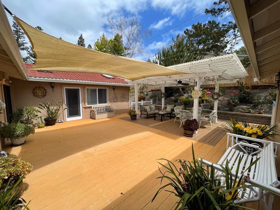 Outdoor patio area with wooden deck flooring, a beige shade sail, and a white pergola with seating arrangements including benches and chairs. The area is decorated with potted plants and flowers, with a garden and trees in the background under a partly cloudy sky.