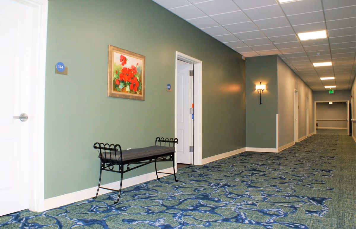 Interior hallway of a senior living facility with doors, a bench, and wall art.