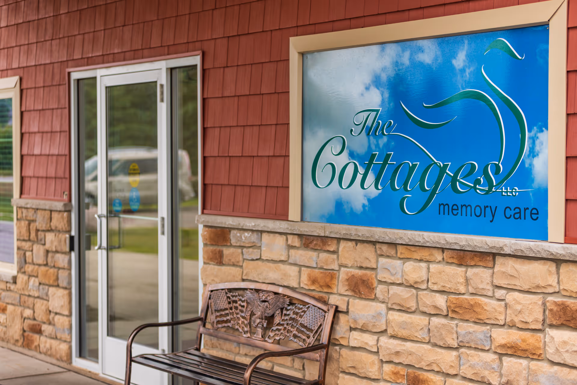 Front entrance of The Cottages memory care facility showing a large blue sign, glass doors, stone facade and a bench.