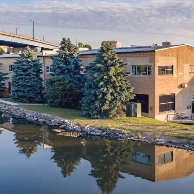 Brick facility by a calm water body with evergreen trees and a stone embankment.