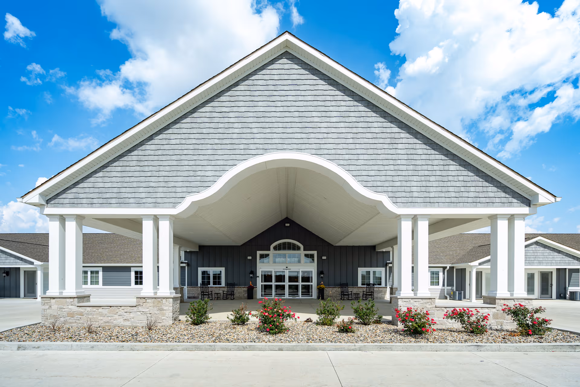 Front entrance of an assisted living facility with a large covered porte-cochere, white columns, and landscaped flower beds.