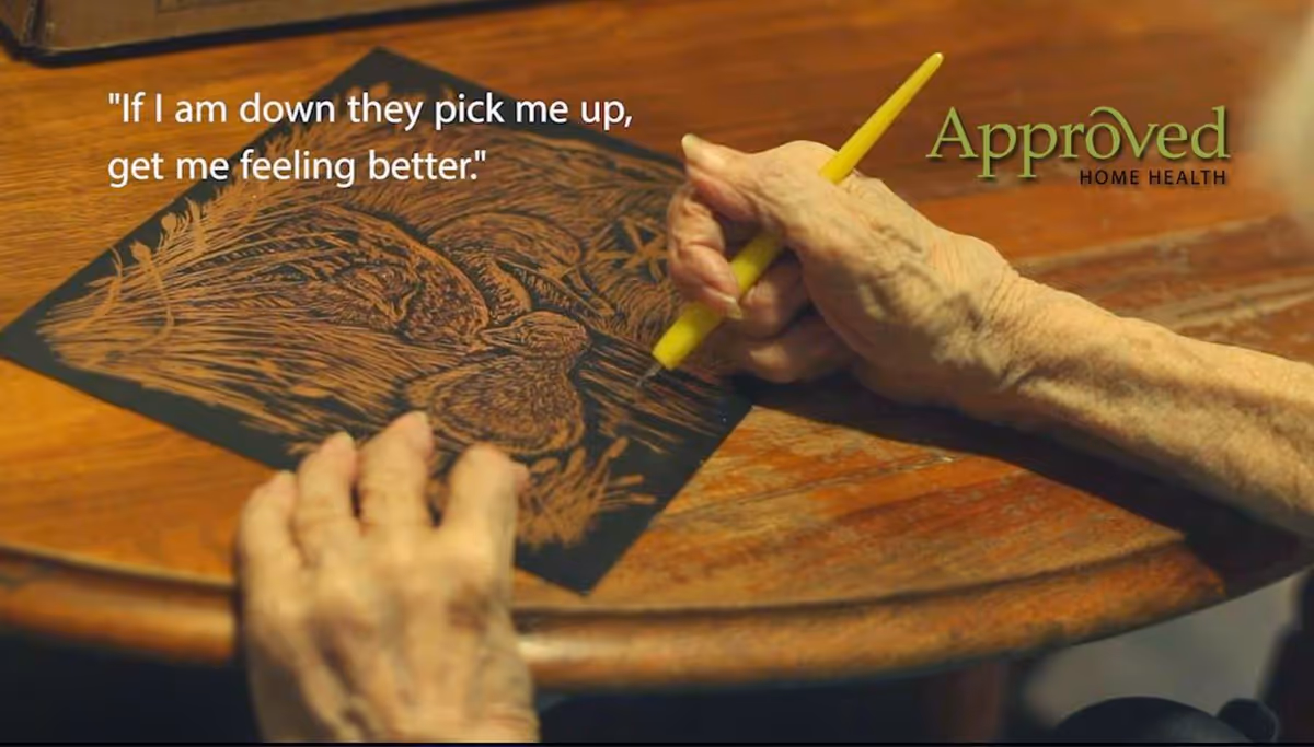 Elderly hands draw on a dark paper with a yellow pencil at a wooden table.