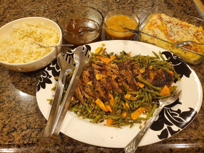 A meal served on a granite countertop featuring a large plate with sliced roasted meat and green beans with carrots, a bowl of white rice, two small glass bowls with sauces, and a glass baking dish with a baked casserole topped with cheese.