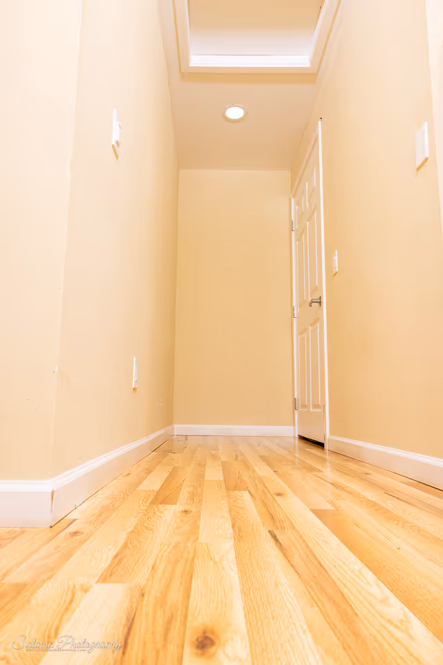 Narrow beige hallway with wooden floor, door on the right, and skylight overhead.
