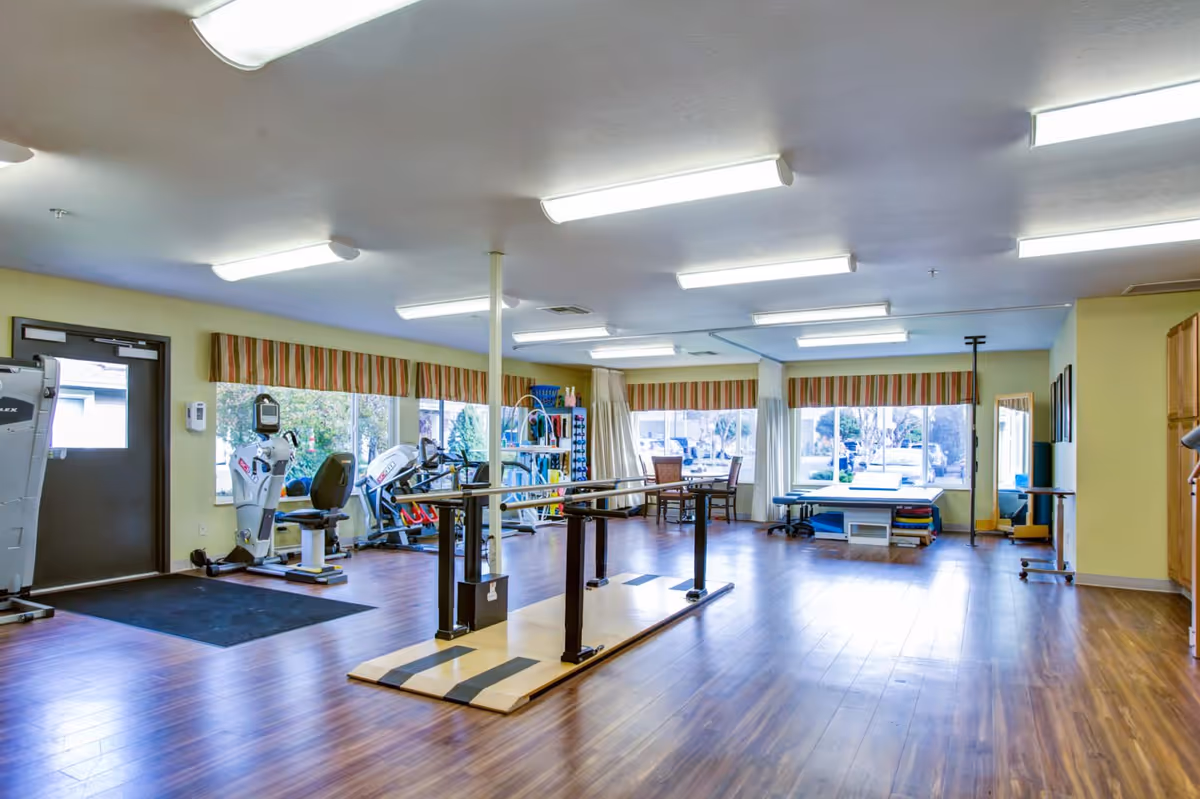 Bright, spacious rehabilitation/exercise room with parallel bars, exercise machines, tables, and large windows.