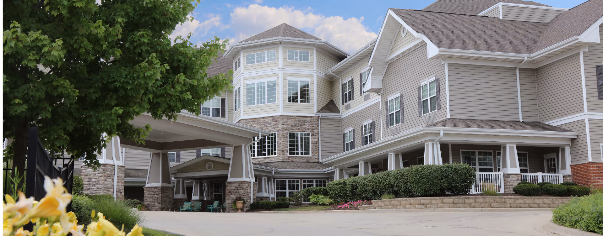 A modern senior living facility exterior with a wide entrance, manicured landscaping, and driveway.