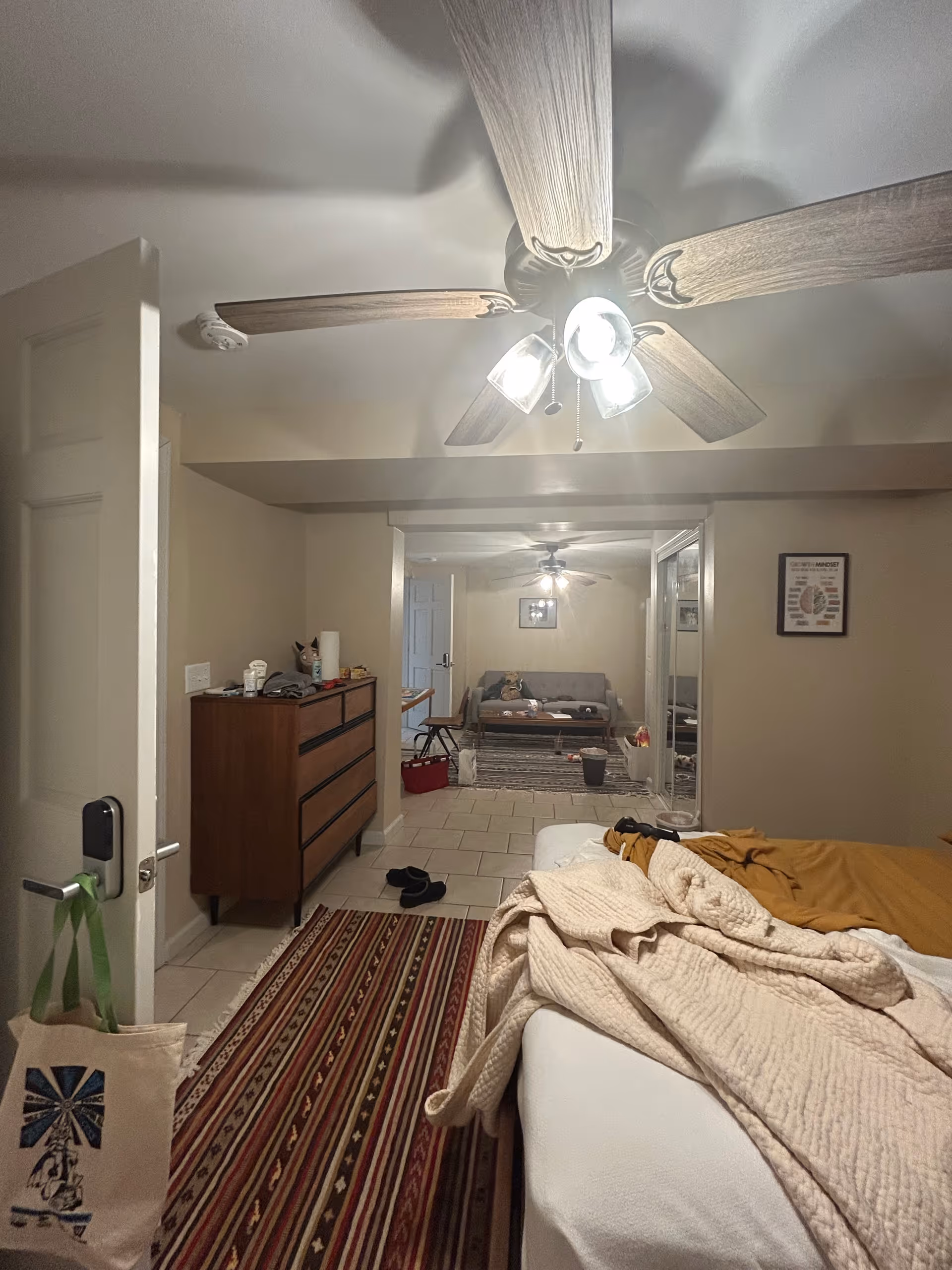 Bedroom in a senior facility with a bed, dresser, and open living area beyond.