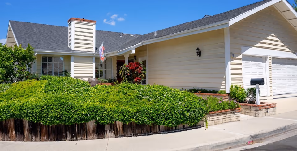 Single-story beige senior living facility with manicured hedges, a driveway, and a white garage.