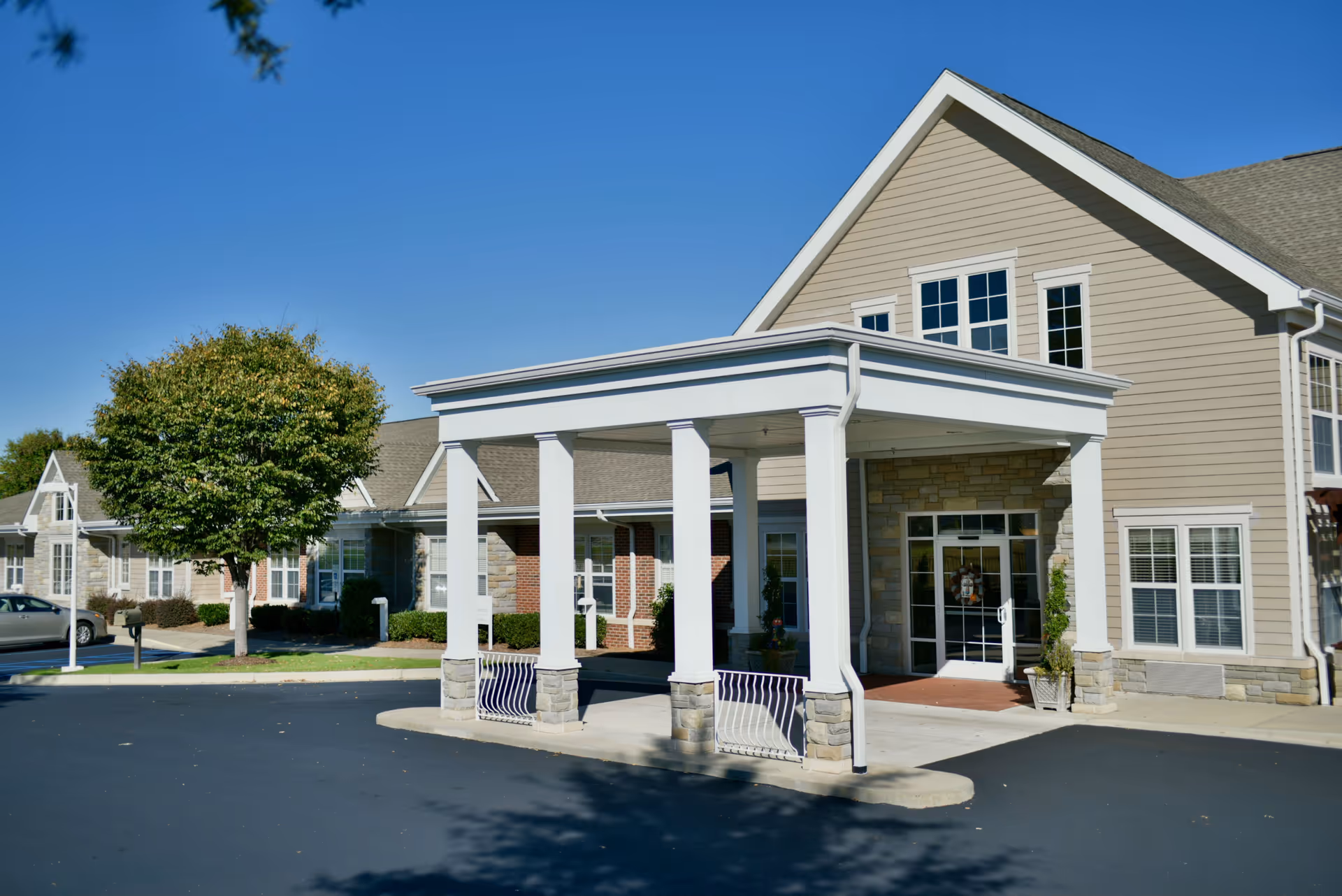Front entrance of a senior living facility with a covered porte-cochere supported by white columns and beige siding.