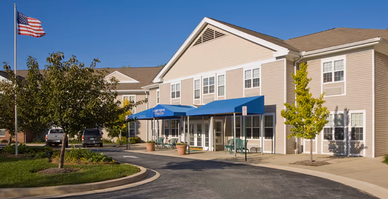 Two-story beige senior living building with blue awnings over the main entrance, flagpole, parked cars, and landscaped driveway.