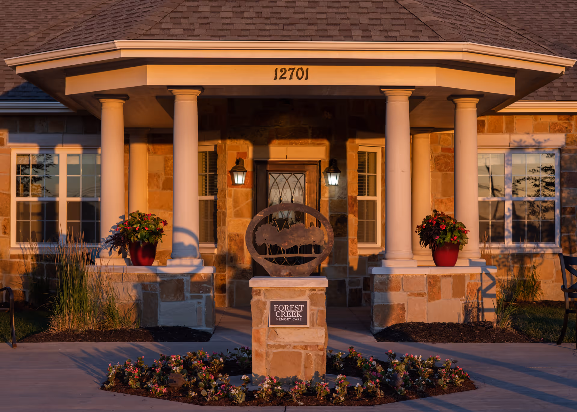Front exterior view of a memory care community building with stone walls, four white columns supporting a porch roof, two red flower pots on stone ledges, and a decorative sculpture on a stone pedestal surrounded by flowers. The building number 12701 is displayed above the porch.