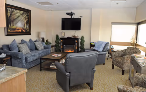 Cozy senior living lounge with blue sofas, armchairs, coffee table, fireplace, and TV.