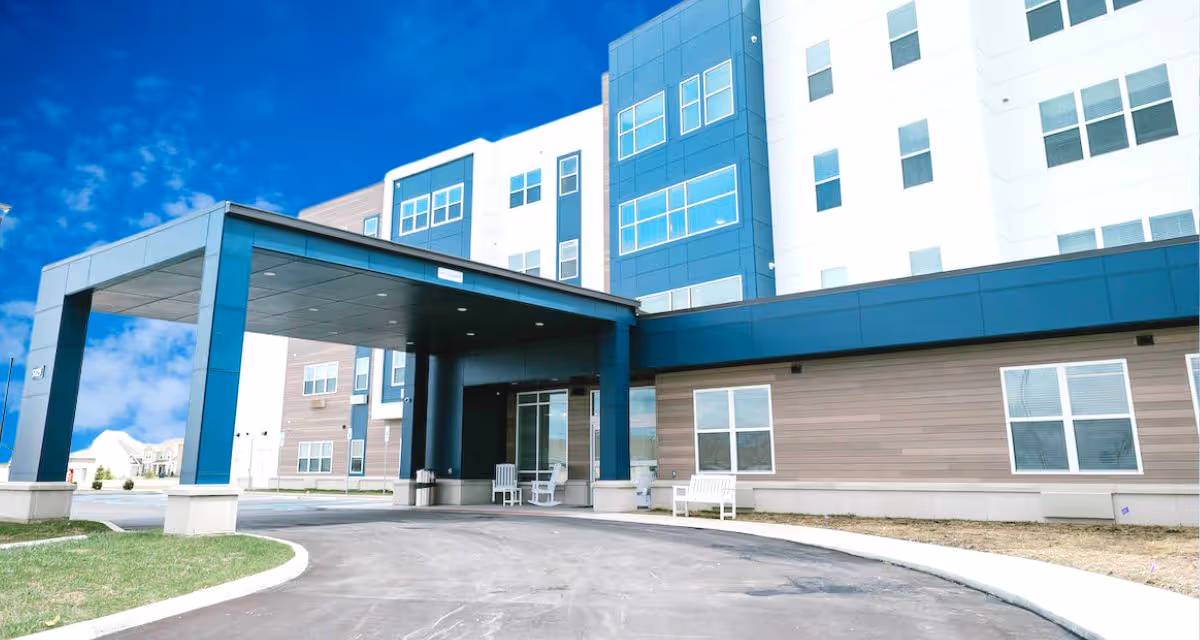 Exterior view of a modern multi-story senior living facility with a covered entrance, large windows, and a driveway leading up to the entrance. There are white benches near the entrance and a clear blue sky in the background.