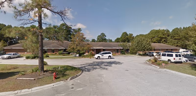 Exterior view of Cedar Cove Assisted Living And Memory Care facility showing a single-story brick building with a parking lot in front, several parked vehicles, trees, and a clear sky.