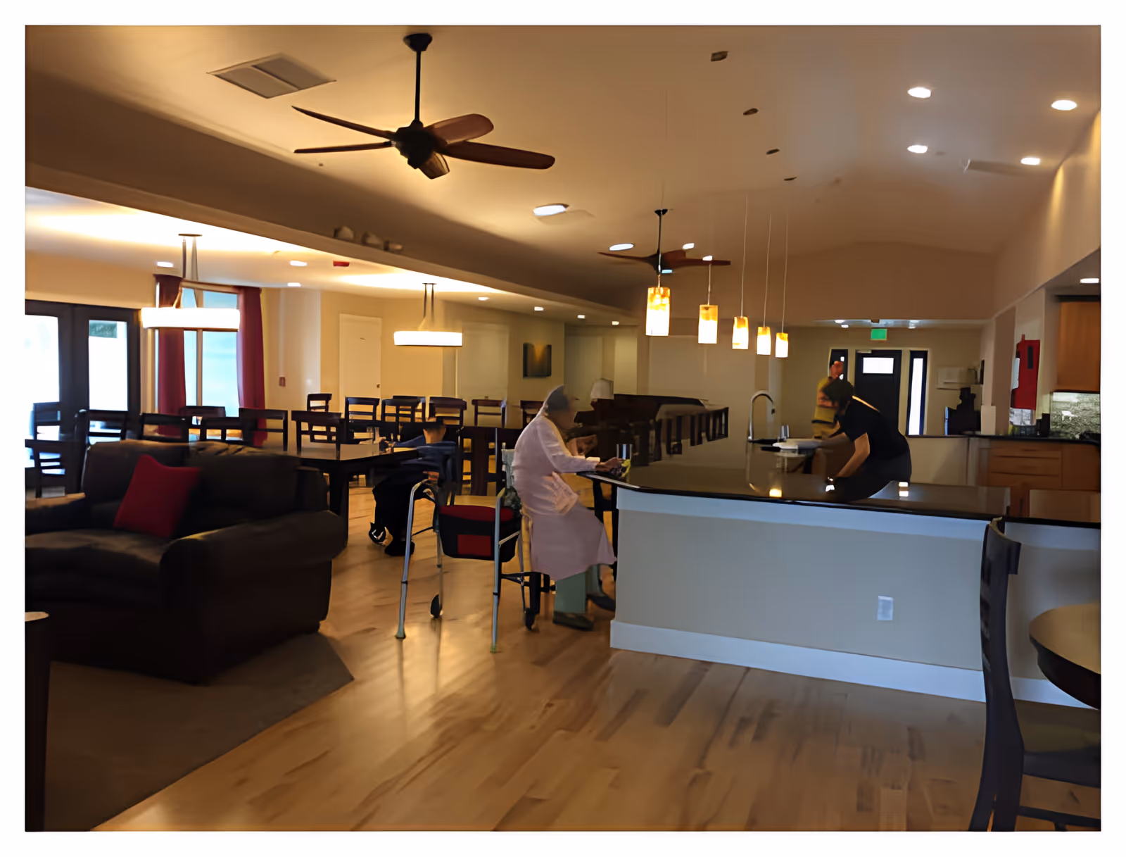 Interior view of a spacious assisted living facility common area with wooden floors, a dark brown couch with red pillows, multiple dining tables and chairs, ceiling fans, and pendant lights hanging over a kitchen island. Two people are present, one sitting at the kitchen island and another standing nearby.