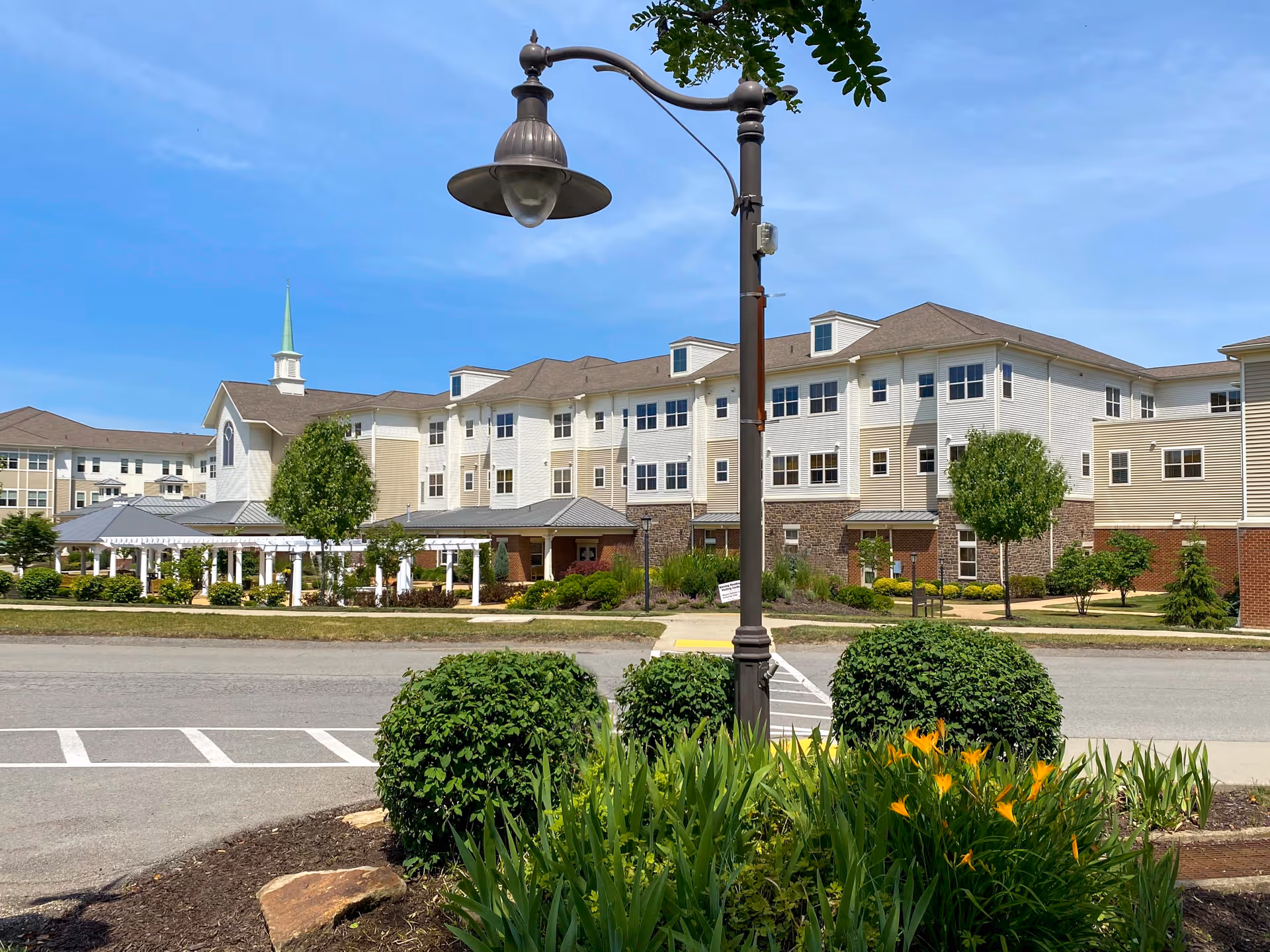 Front exterior of a multi-story senior living facility with a central lamppost, landscaped shrubs, and a covered walkway.