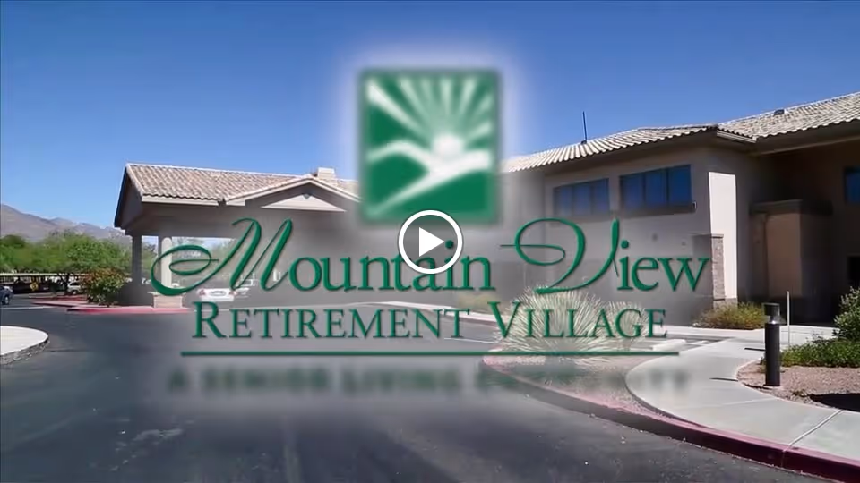 Front exterior and entrance of a retirement community building with a driveway and a large overlaid "Mountain View Retirement Village" logo.