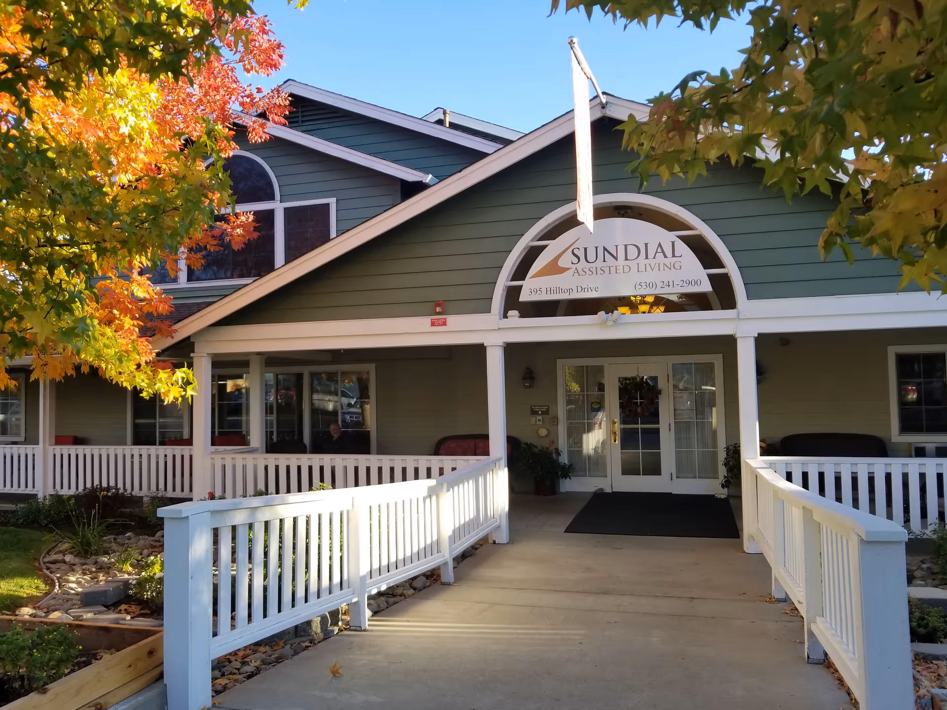 Exterior front entrance of Sundial Assisted Living facility with a green building, white railing along a concrete walkway, autumn trees with colorful leaves, and a sign above the door displaying the facility name and contact information.