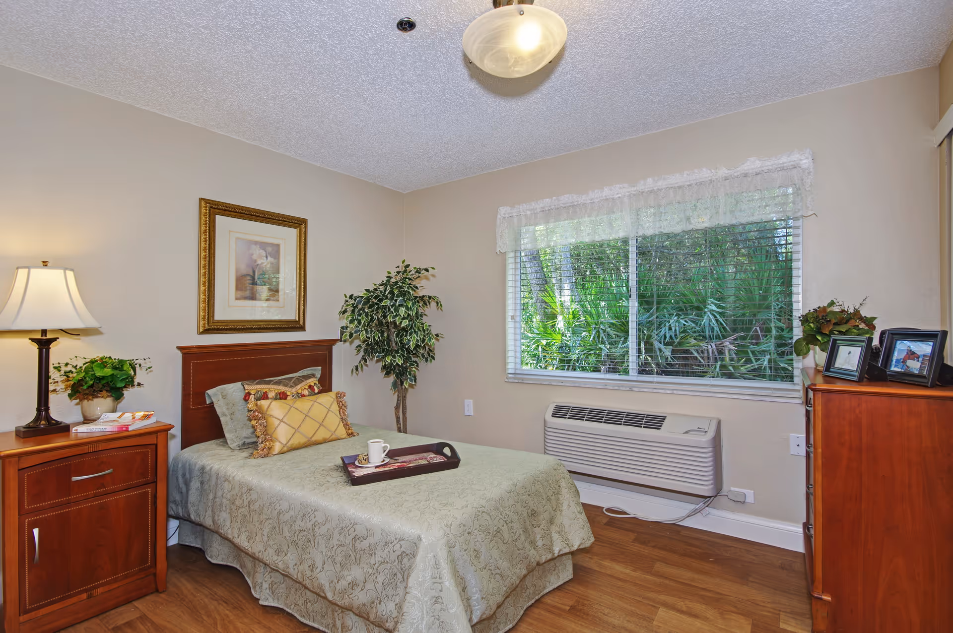 Bedroom in a senior living facility with a bed, furniture, plants, and a window view.