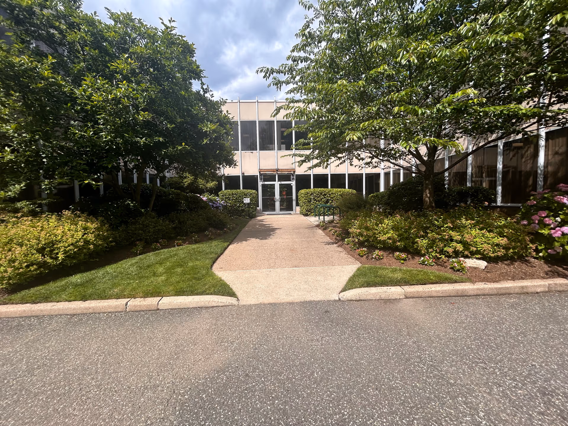 Front entrance of a senior living facility with glass doors, landscaped walkway, and trees.