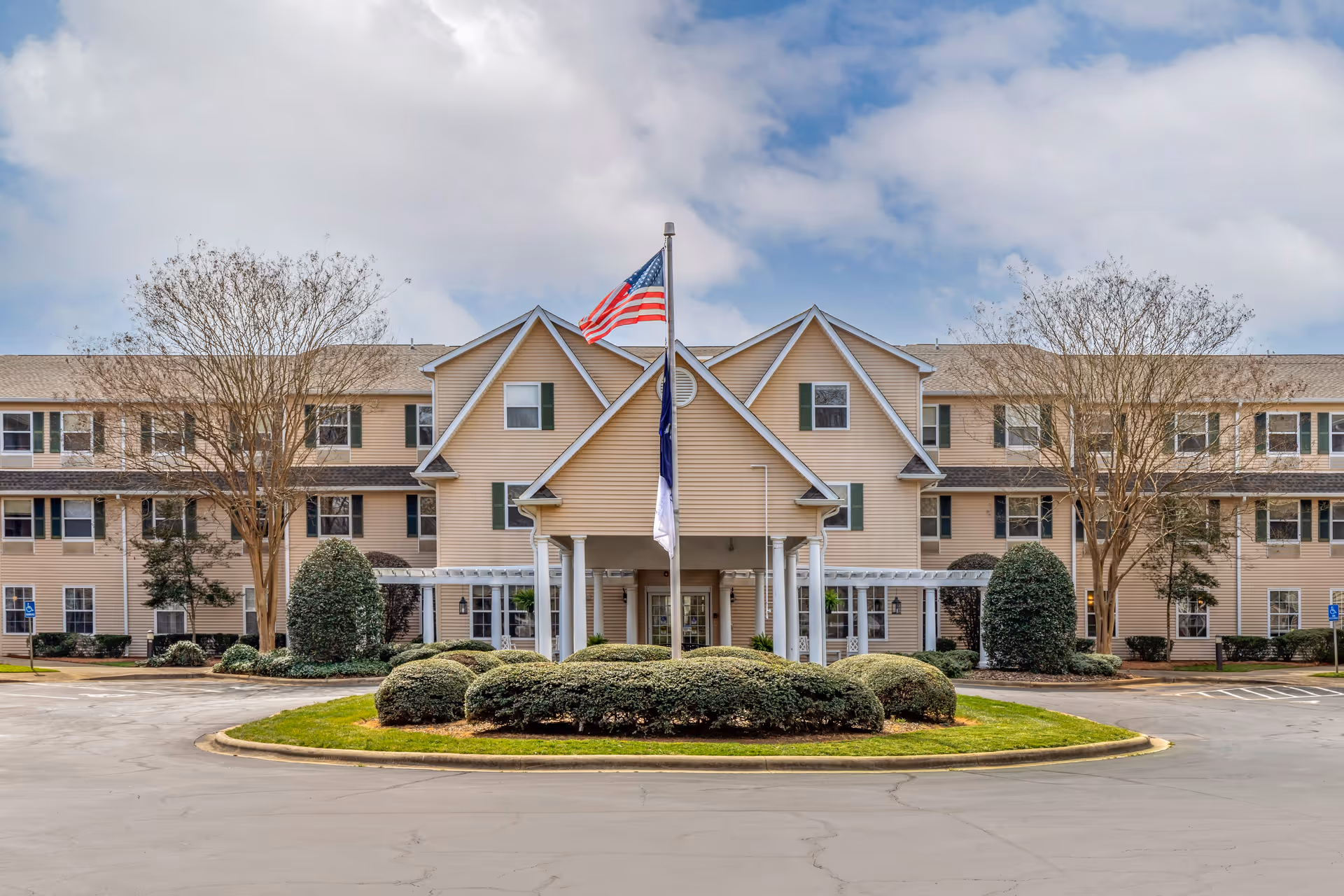 Front exterior view of a three-story assisted living and memory care facility with beige siding, green shutters, and a covered entrance. There are two flagpoles with flags in front of the building, surrounded by neatly trimmed bushes and trees. The sky is partly cloudy.