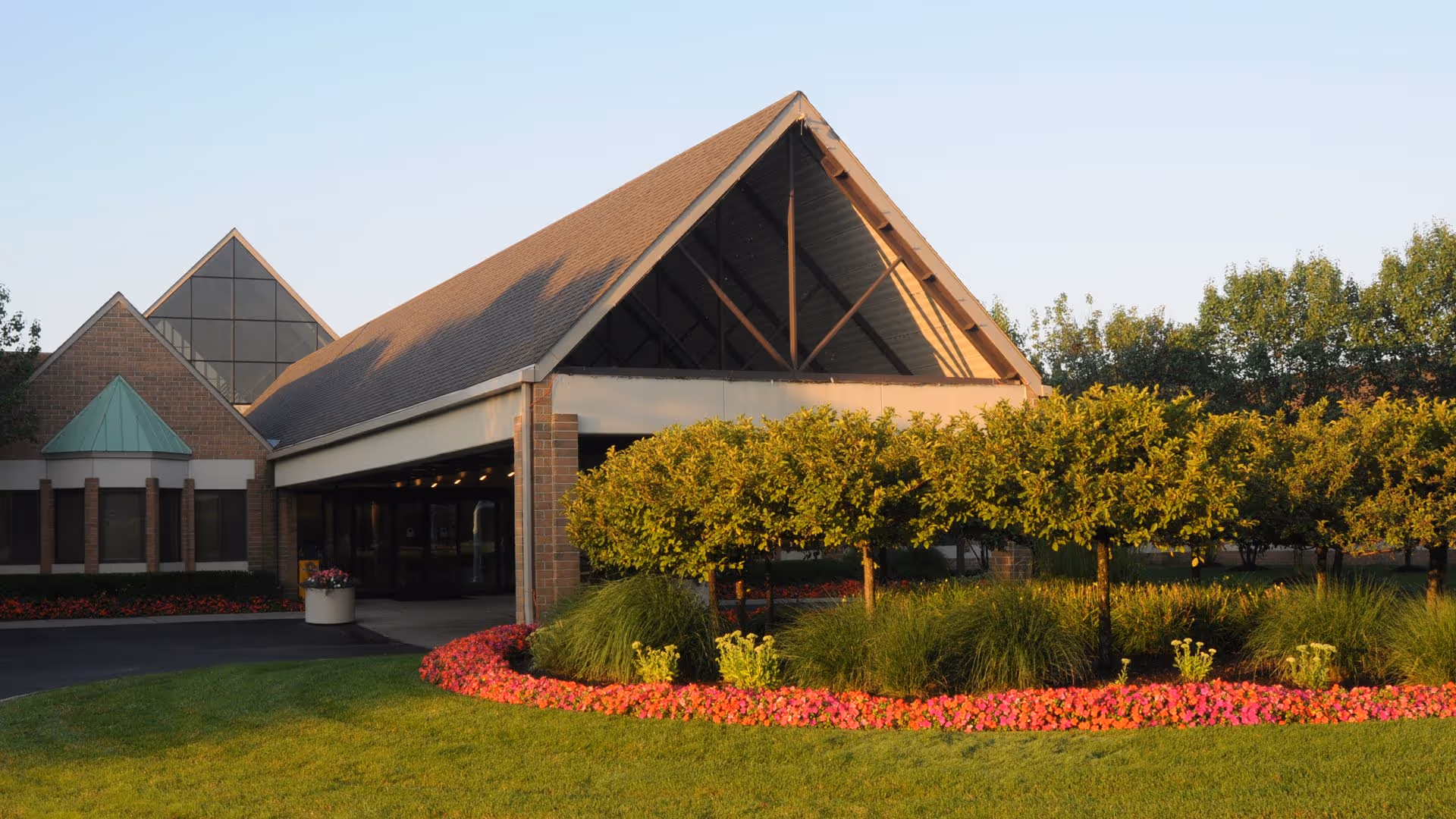 Front entrance of a senior living facility with a peaked canopy, driveway, and manicured flower beds and trees.