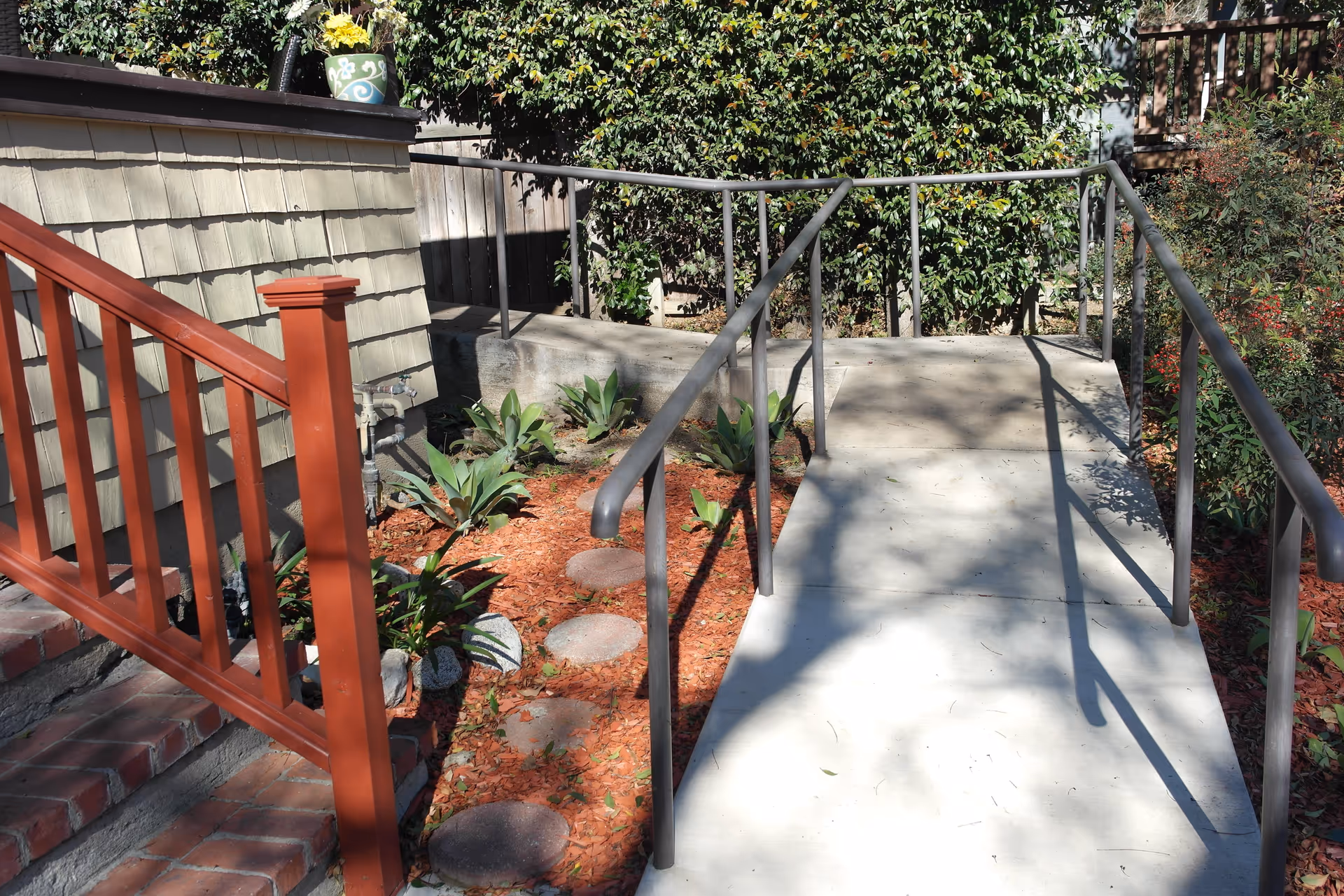 Concrete wheelchair ramp with metal handrails alongside brick steps and landscaped plant beds at a house exterior.