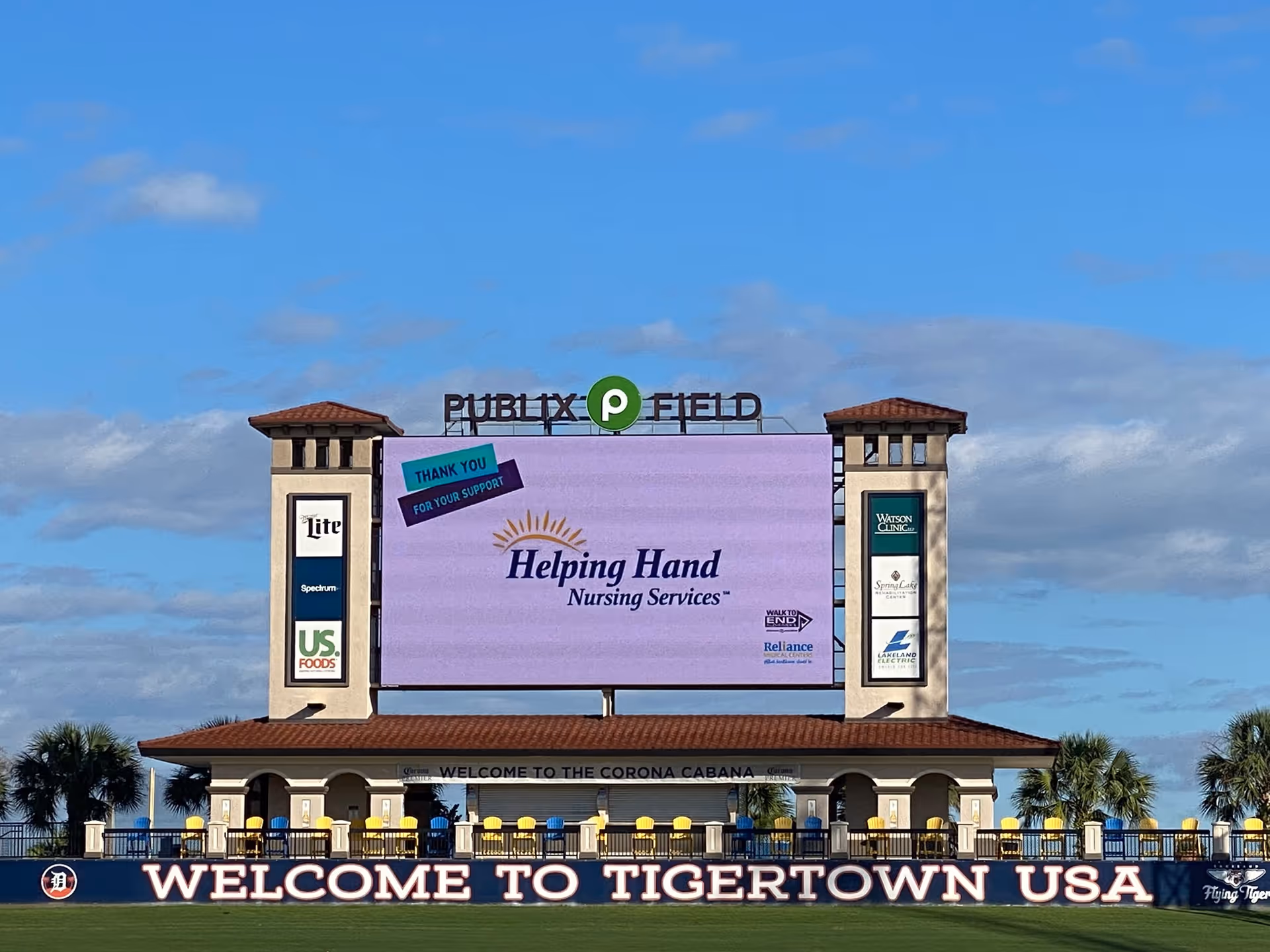 Outdoor stadium with a large screen displaying Helping Hand Nursing Services on a purple billboard.