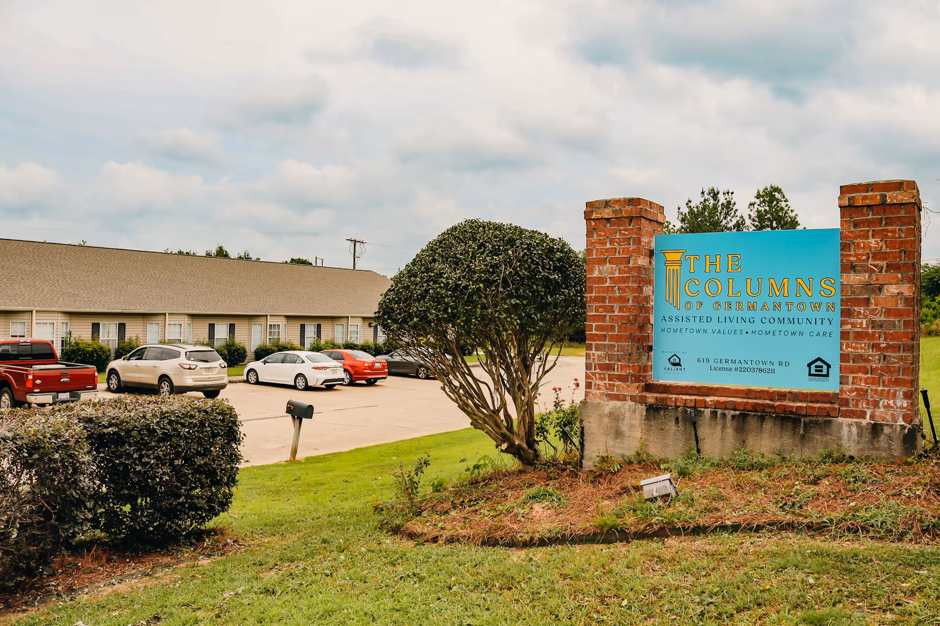 Exterior view of The Columns of Germantown Assisted Living Community sign with a brick frame, a parking lot with several cars, and a single-story building in the background under a cloudy sky.