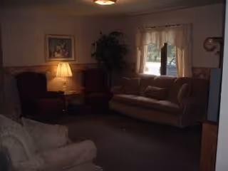 Dimly lit living room with beige sofa, armchairs, lamp, plant, and window curtains.