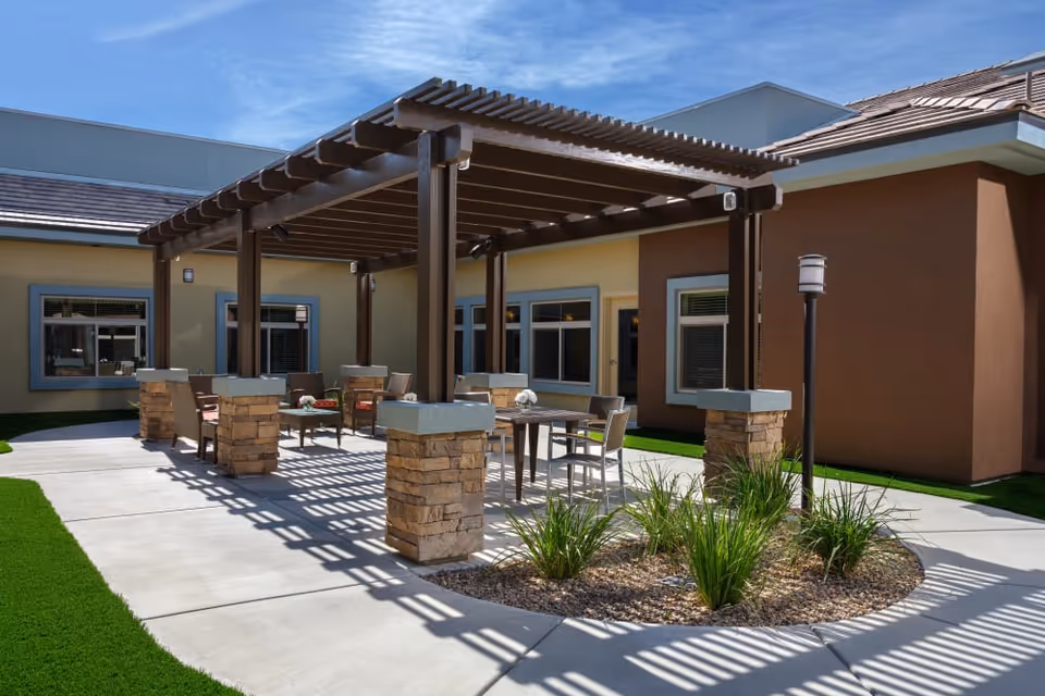 Outdoor courtyard patio with a shaded pergola, tables and chairs, stone columns and landscaping outside a senior living facility.