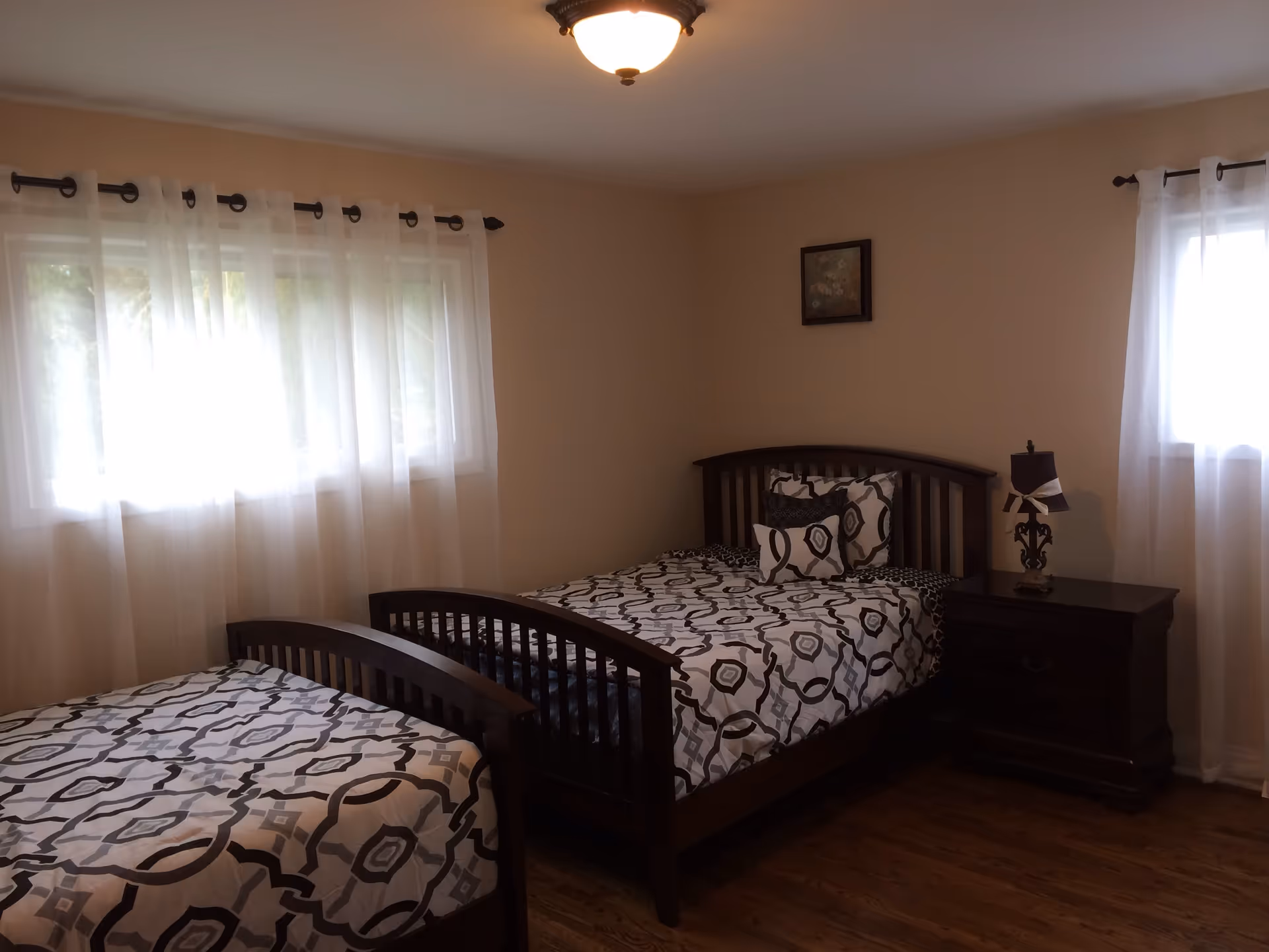 A bright bedroom with two wooden twin beds with patterned bedding, a nightstand with a lamp, and sheer curtains over the windows.