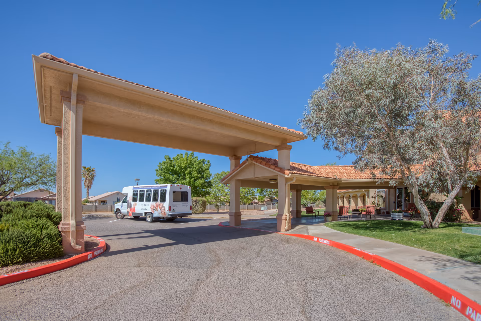 Entrance area of Vista Pointe at Sierra Vista senior living facility with a covered drop-off zone, a shuttle bus parked nearby, and landscaped greenery including trees and bushes under a clear blue sky.