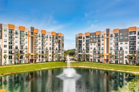 Symmetrical modern buildings frame a central pond with a fountain and palm trees.