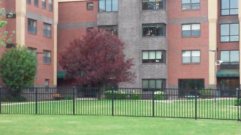 Multi-story brick building with green lawn, trees, and a black metal fence in front.