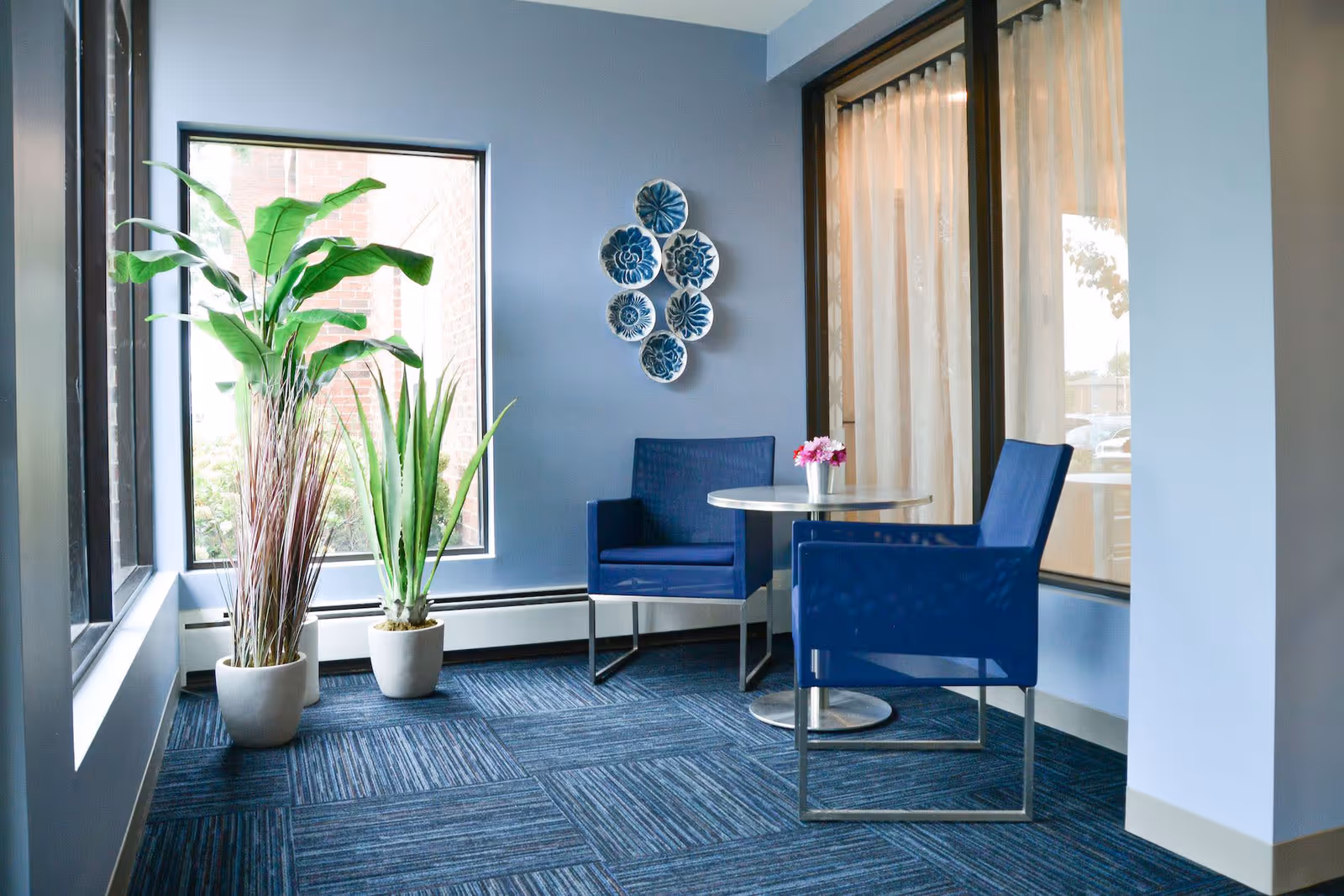 A small seating area with two blue chairs and a round table with a small flower pot on it, situated next to large windows with beige curtains. Two potted plants are placed near the windows, and decorative blue and white plates are mounted on the light blue wall.
