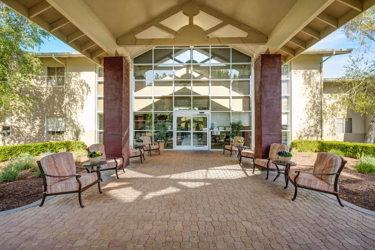 Entrance area of Creston Village facility with a covered walkway featuring cushioned chairs and small tables on both sides. The building has large glass windows and double glass doors, with greenery and trees surrounding the paved pathway.