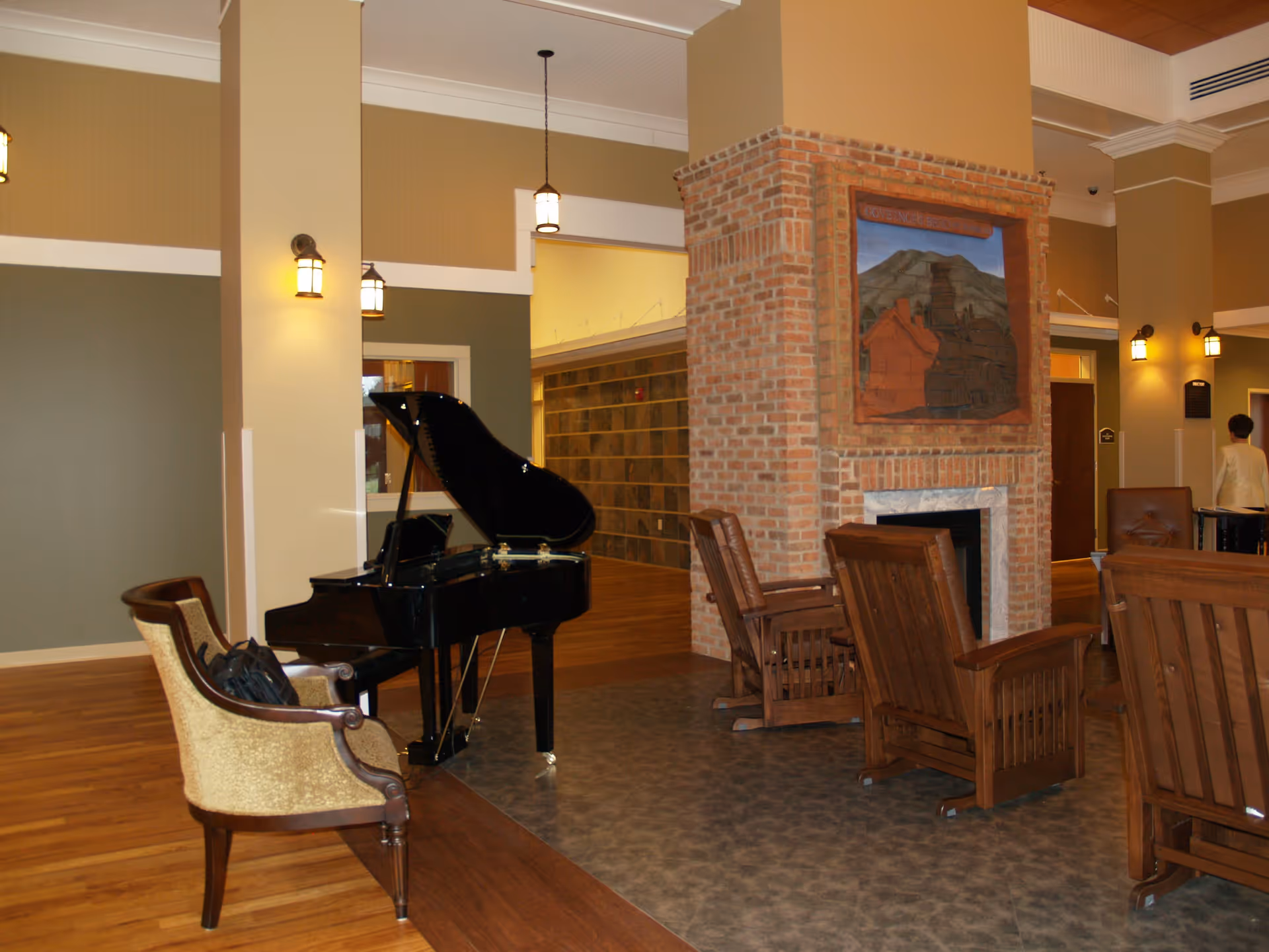 Interior common area of Governor’s Bend Retirement & Assisted Living featuring a black grand piano, a beige upholstered chair, wooden armchairs arranged around a brick fireplace with a decorative tile mural above it, and warm lighting fixtures on the walls and ceiling.