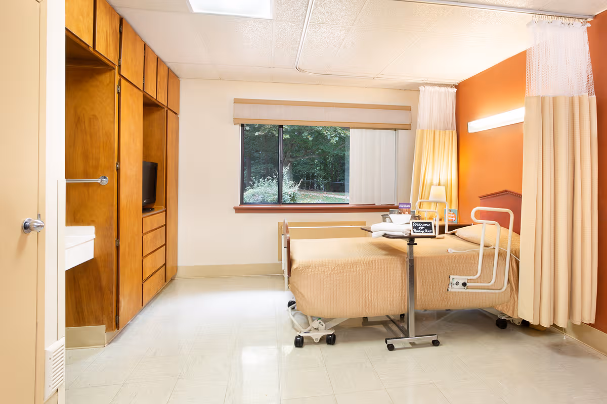Senior living patient room with single bed, curtains, dresser, wall-mounted light, and window view.