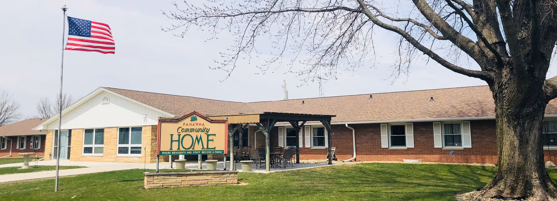 Single-story brick senior living facility with lawn, flagpole, sign, and outdoor seating area.