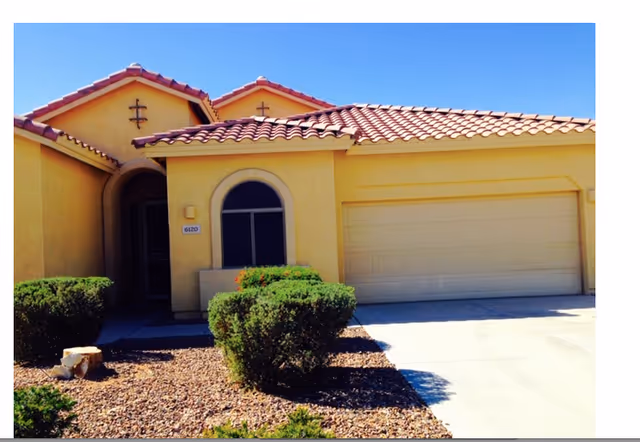 Exterior front view of a single-story yellow stucco house with a red tile roof, a garage door, an arched window, and a small landscaped area with bushes and rocks in front.
