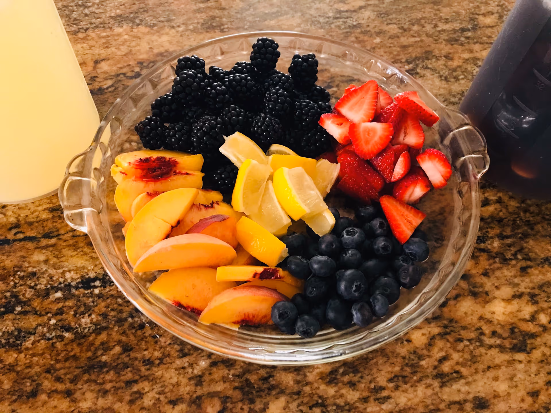 Glass bowl of assorted fruit (blackberries, strawberries, blueberries, peach slices and lemon wedges) on a granite countertop.