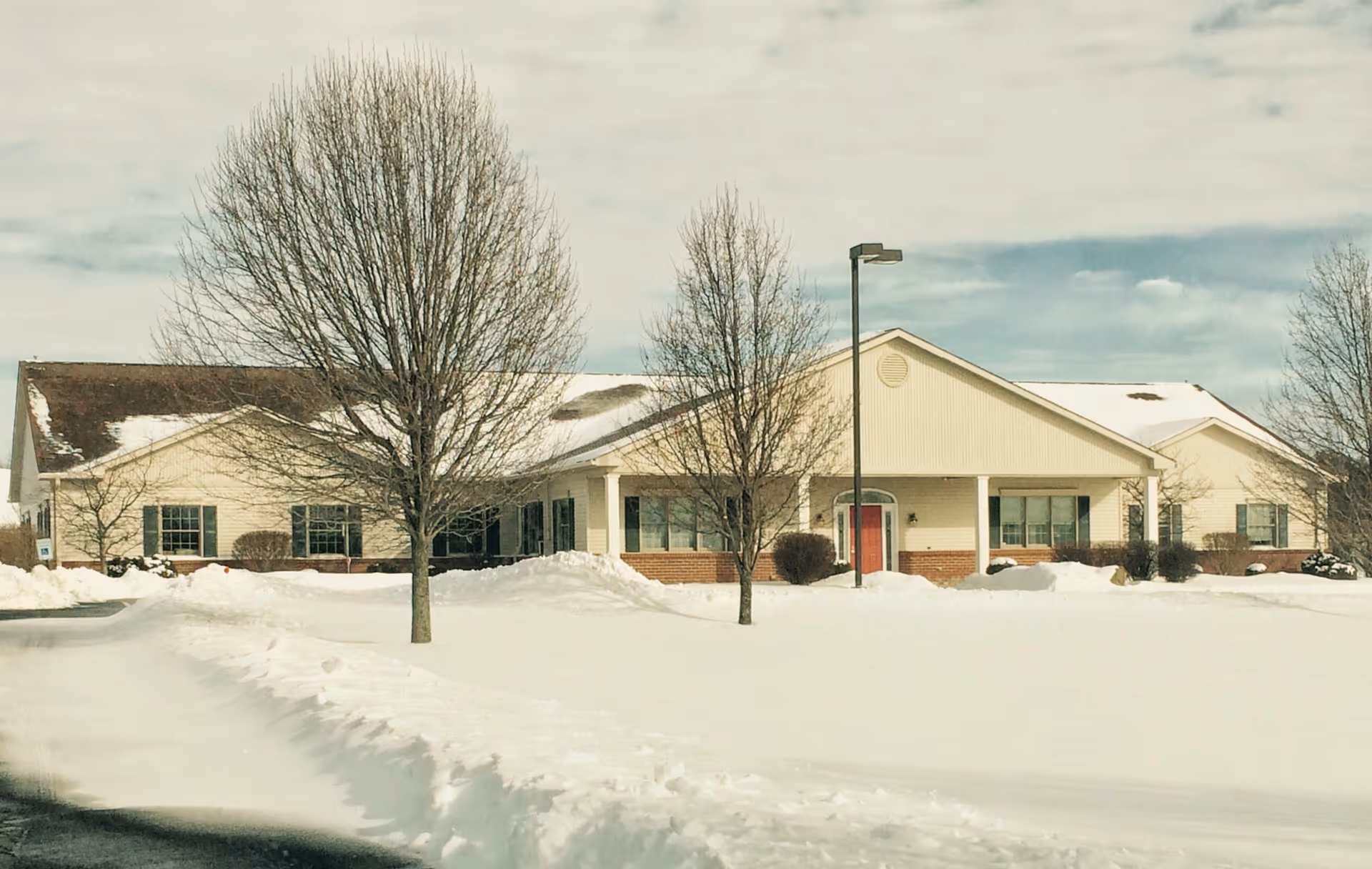Snow-covered exterior of a senior living facility with entrance and leafless trees.